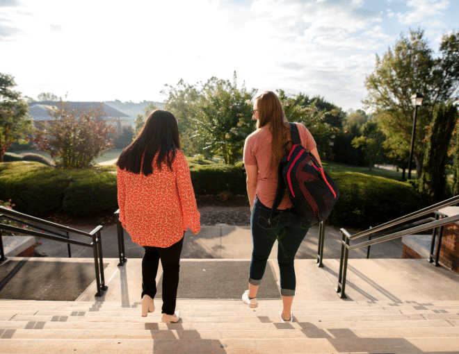 Two people walk down sunlit outdoor steps, one wearing a red patterned top and the other carrying a backpack. Green trees and bushes line the background under a partly cloudy sky.