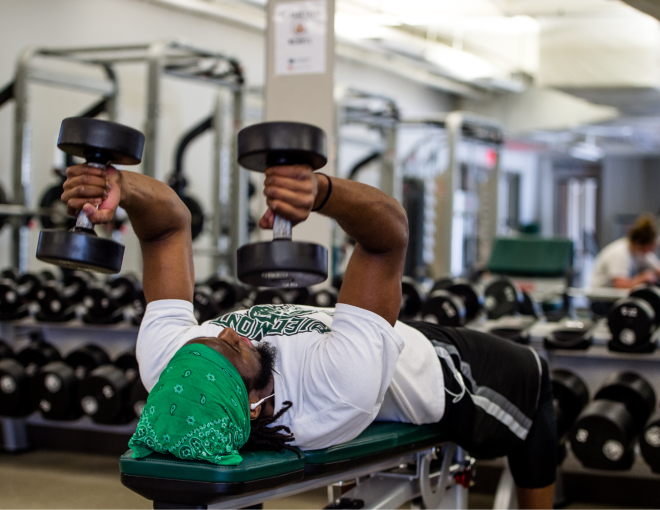 A person wearing a green bandana and white shirt is lying on a bench in a gym, lifting two dumbbells above their chest. Rows of dumbbells and gym equipment are visible in the background.
