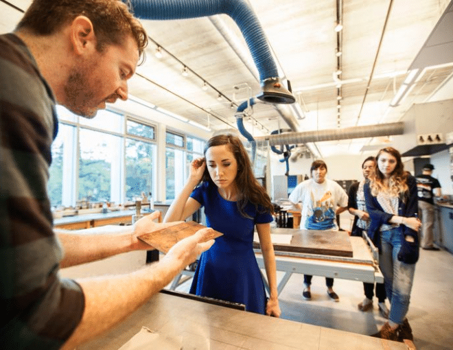 A man shows a piece of material to a woman in a blue dress in a bright workshop, while three other people observe in the background. The room features large windows and industrial equipment.