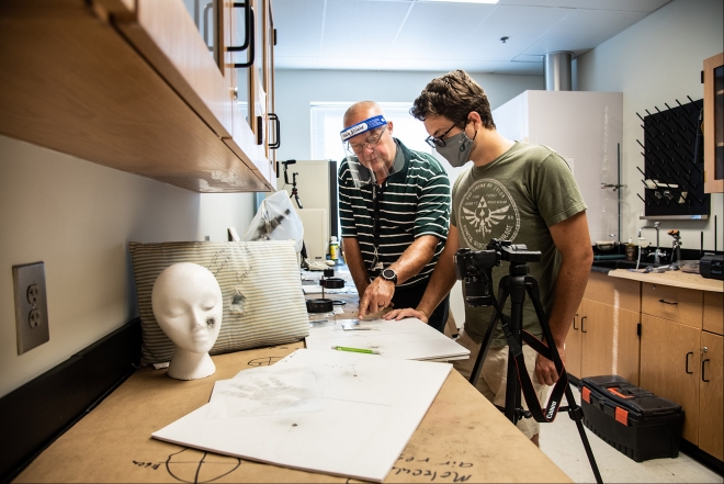 Two people wearing face shields and masks examine papers on a lab table. A mannequin head, camera tripod, and scientific equipment are visible in the laboratory setting.