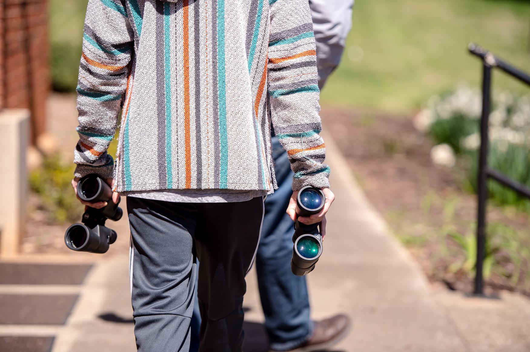 Student wearing a striped long sleeved shirt, carrying two pairs of binoculars.