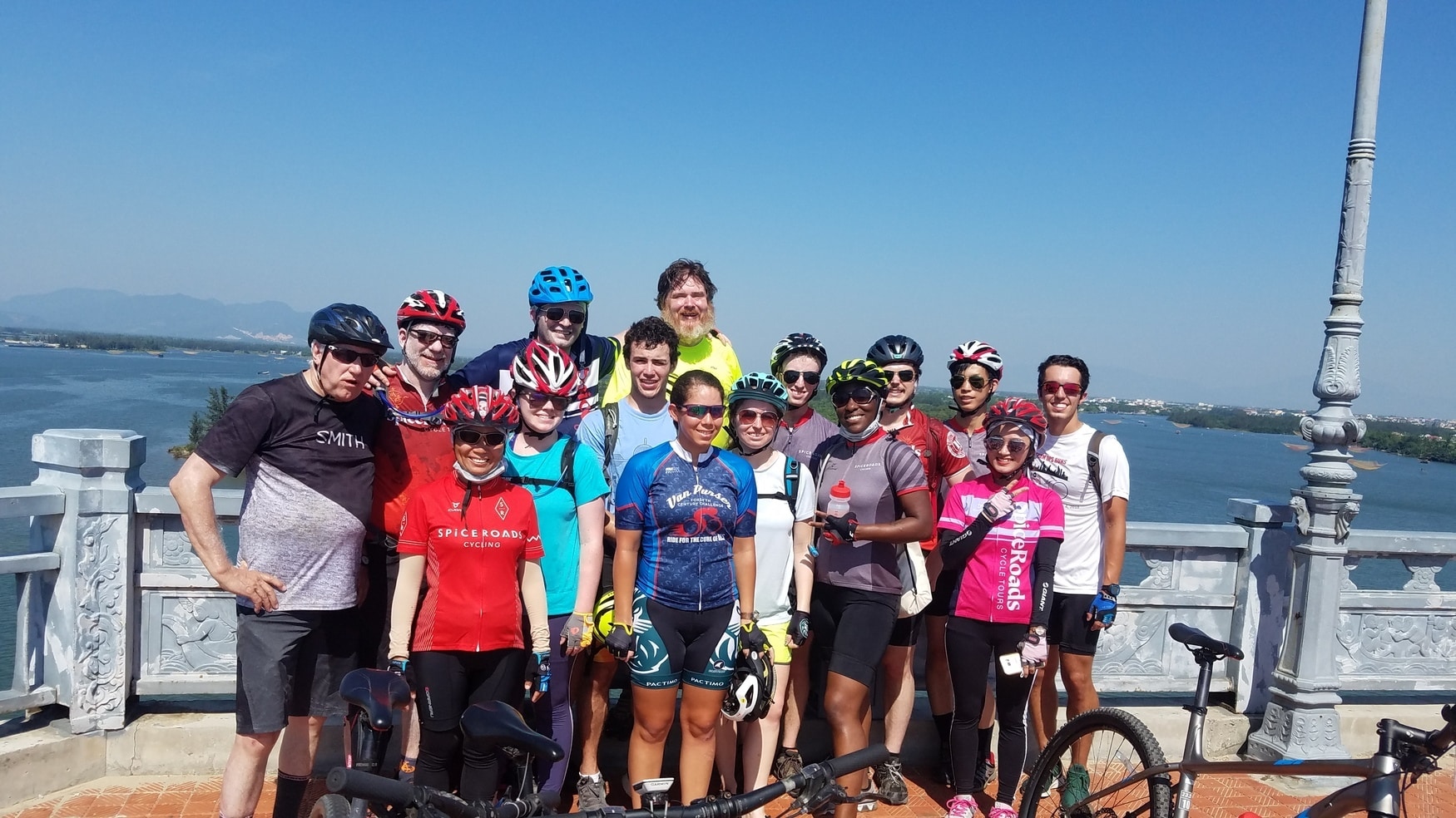 International studies students wearing bicycle gear, standing on a bridge and posing for a picture. A river and mountains are visible in the background.