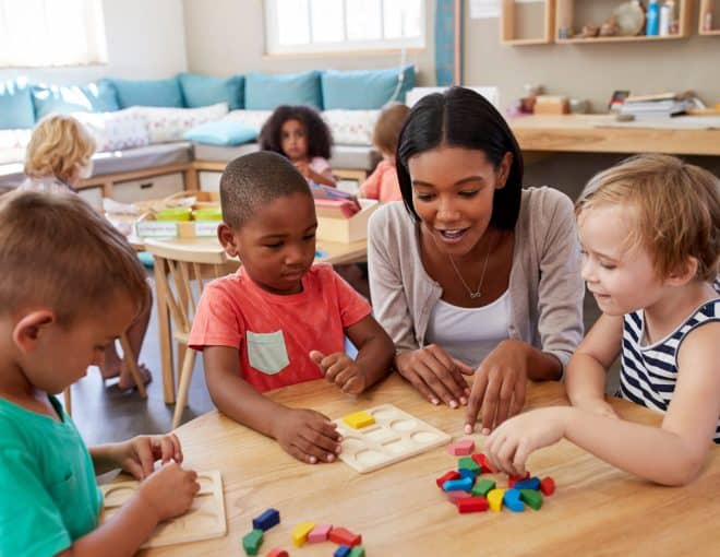 A teacher sits at a table with three young children, helping them with educational toys and puzzles in a bright, cheerful classroom. Other children play in the background.