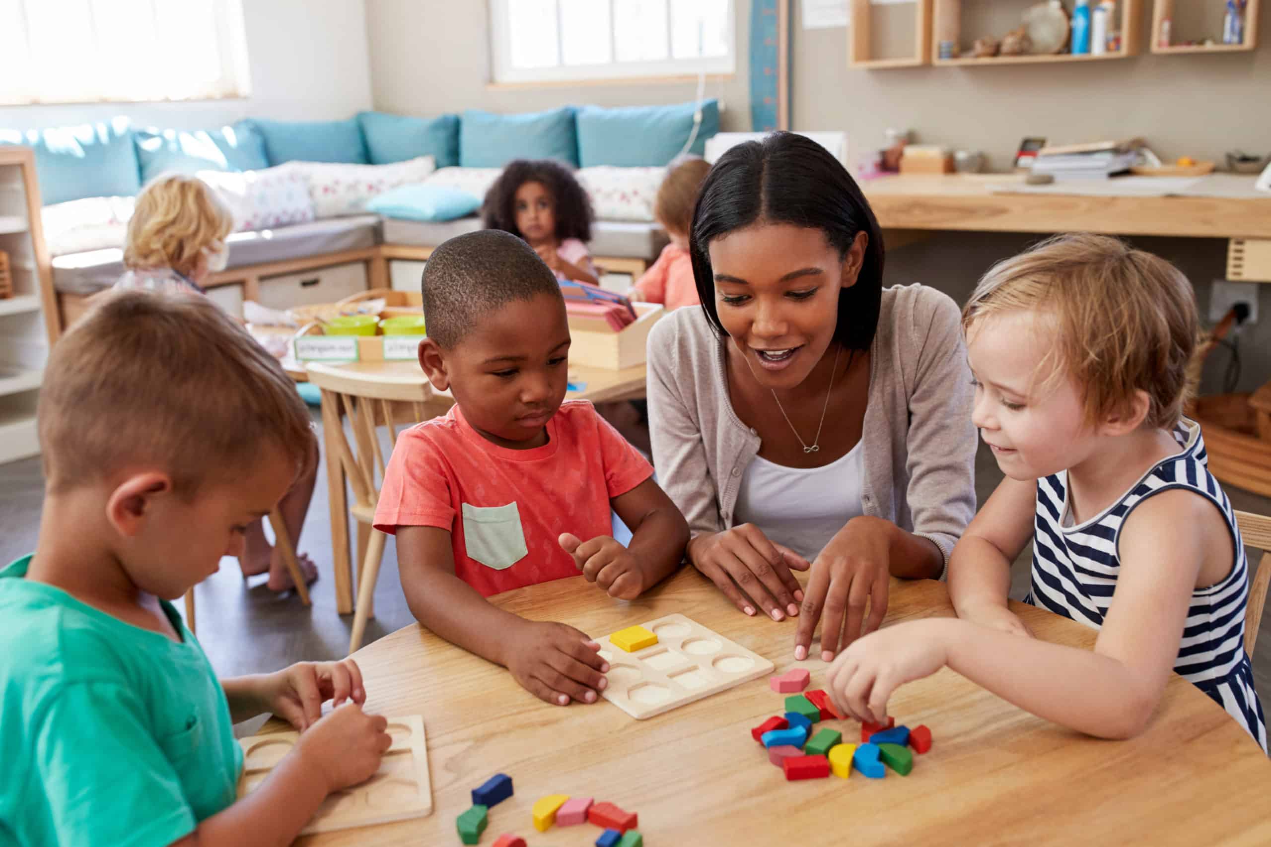 A teacher sits at a table with three young children, helping them with educational toys and puzzles in a bright, cheerful classroom. Other children play in the background.