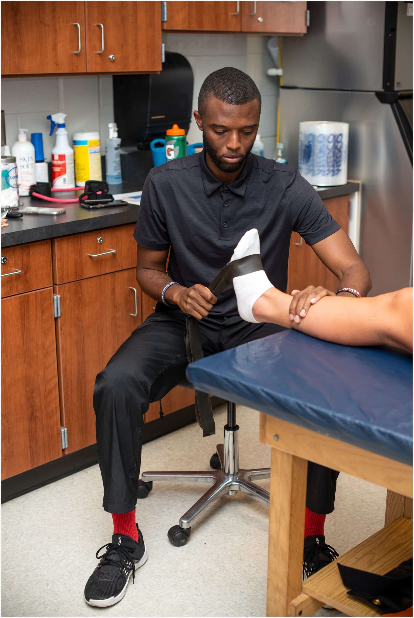 A man in a black shirt and red socks sits on a stool in a medical room, holding and examining the foot of a person lying on an exam table, whose leg is wrapped in a white bandage.