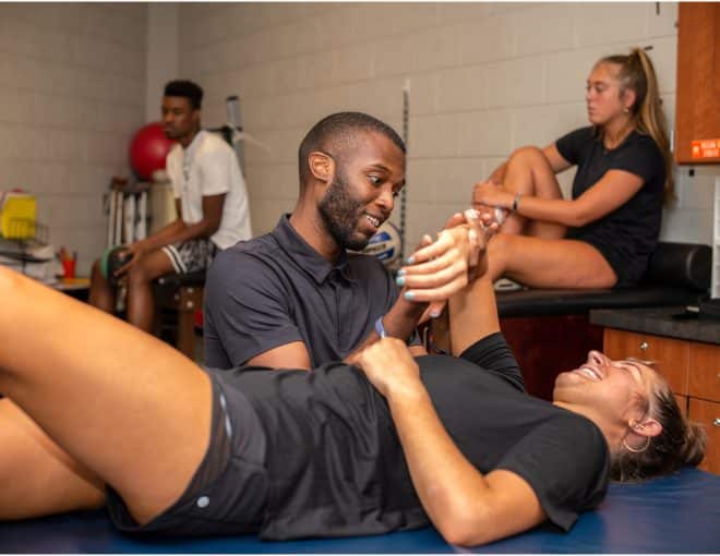 A physical therapist assists a smiling woman lying on a table with arm exercises, while two other people sit and stretch in the background of a sports therapy room.