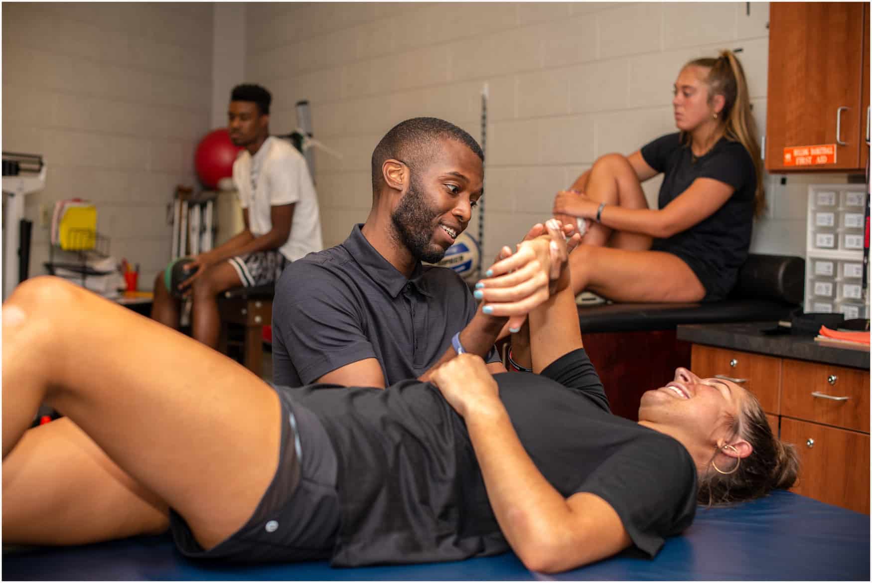 A physical therapist assists a smiling woman lying on a table with arm exercises, while two other people sit and stretch in the background of a sports therapy room.