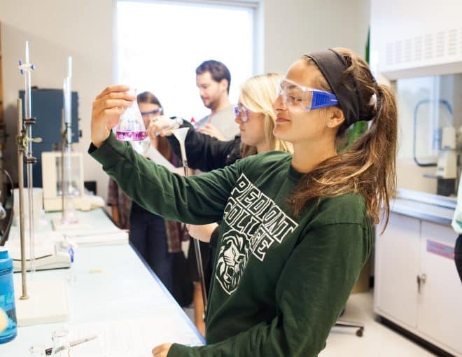 Group of students in a chemistry lab. The focal point is a young woman wearing safety glasses, smiling while holding a beaker full of purple liquid.