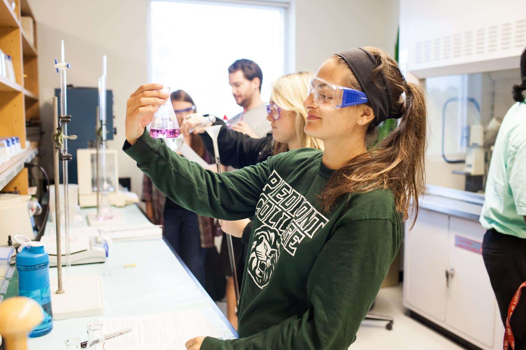 Group of students in a chemistry lab. The focal point is a young woman wearing safety glasses, smiling while holding a beaker full of purple liquid.