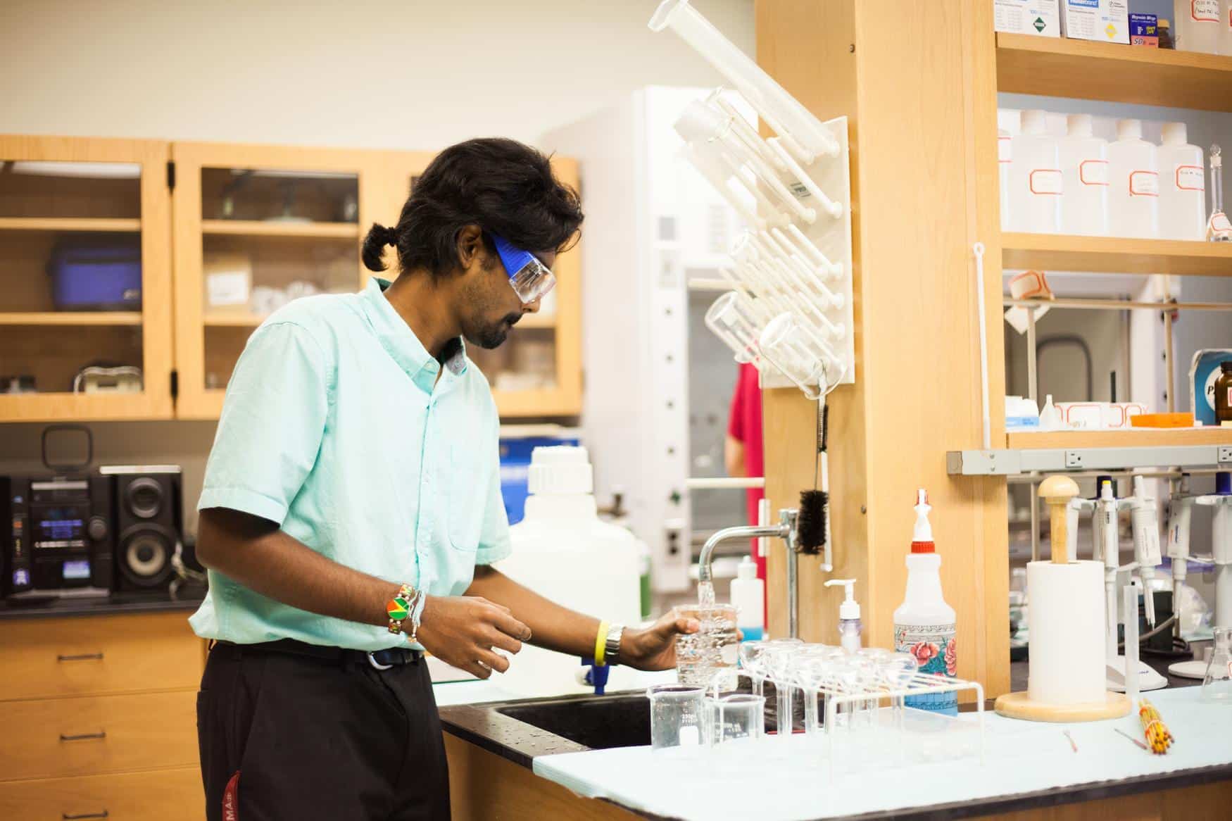 Young man in a laboratory setting with a mint green shirt and safety glasses, filling a beaker full of water in the sink.