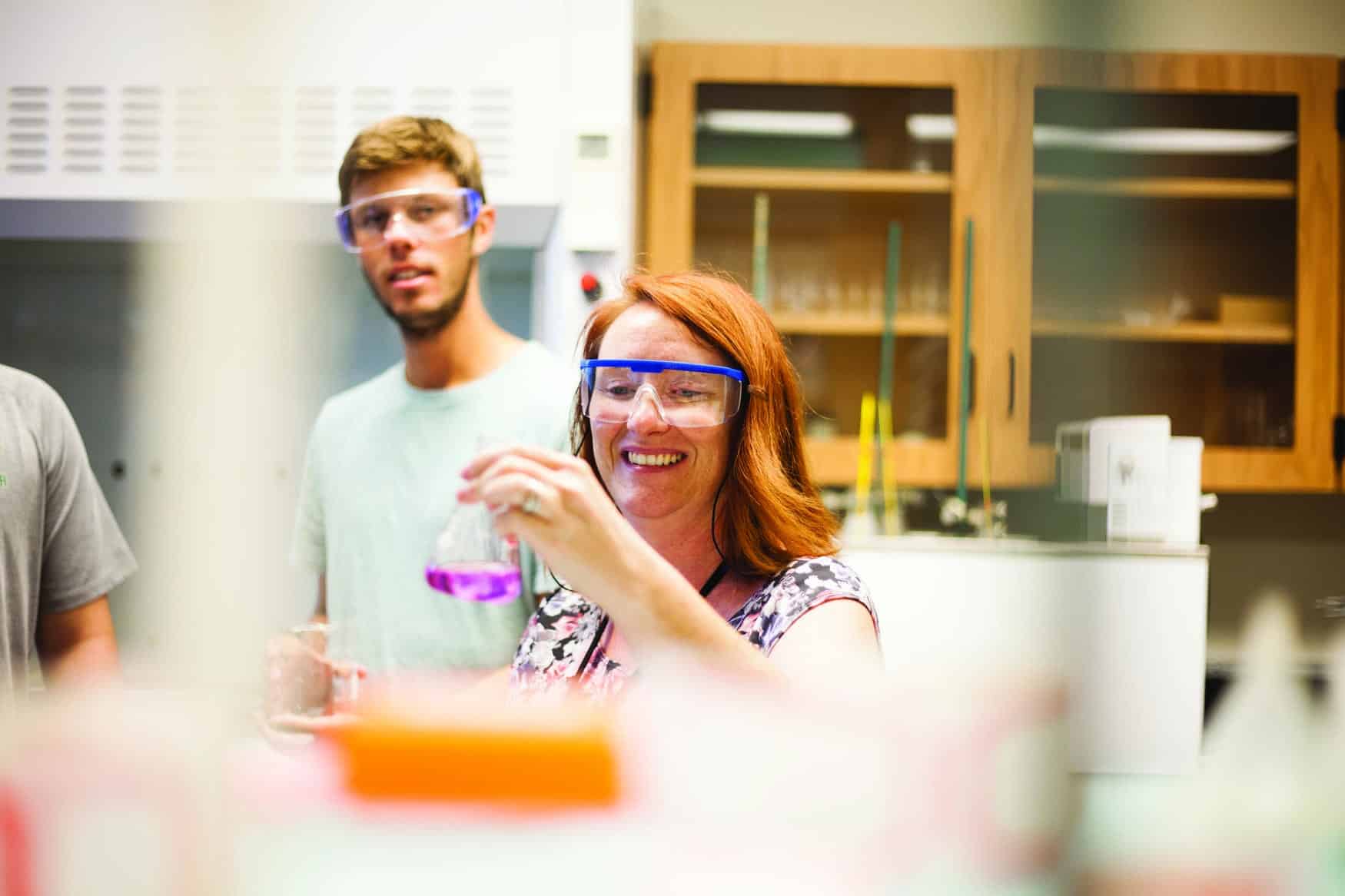 Woman with red hair holding a beaker full of purple liquid. A young man with safety glasses watches in the background.