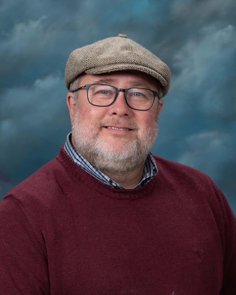 A bearded man wearing glasses, a flat cap, a red sweater, and a plaid collared shirt smiles in front of a blue and gray cloudy backdrop.