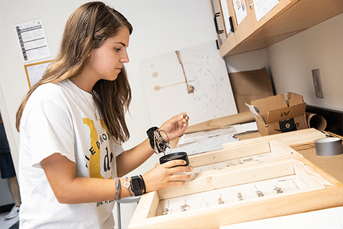 A young woman with long brown hair works at a desk, handling small components and a container, surrounded by wooden frames, tools, and papers in a well-lit workspace.