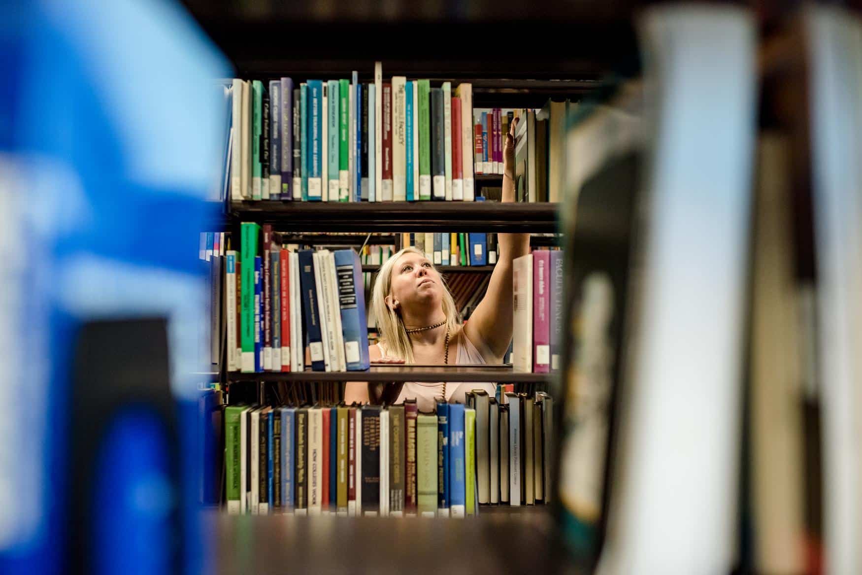 Woman with blonde hair visible through book shelves. She is reaching up to grab a book on a top shelf.