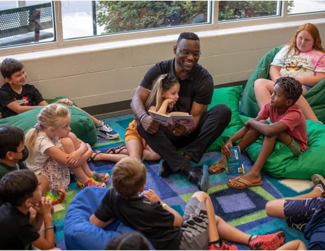 Adult man sitting in a circle with young children. He is reading a book to the children, and the children are listening intently while sitting on green bean bags.
