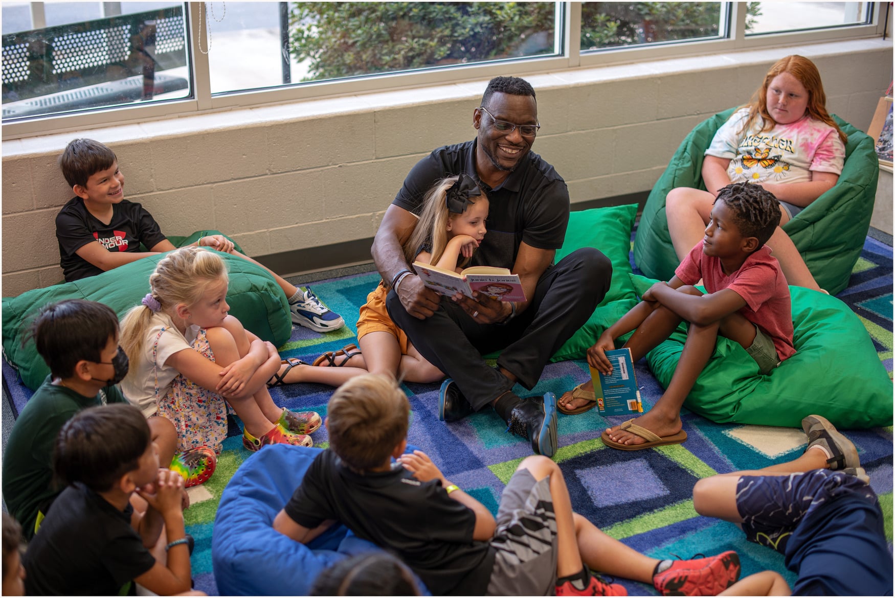 Adult man sitting in a circle with young children. He is reading a book to the children, and the children are listening intently while sitting on green bean bags.