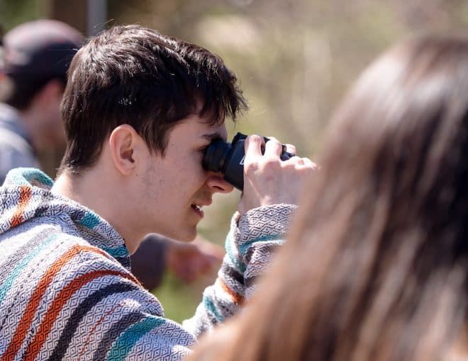 A group of people outdoors, focusing on a young person in a striped hoodie using binoculars. Other individuals nearby are also looking into the distance, possibly birdwatching or observing nature.