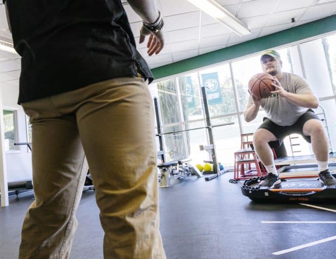 A person squats on a mini trampoline while holding a basketball, facing another person in a gym with bright windows, weights, and exercise equipment in the background.