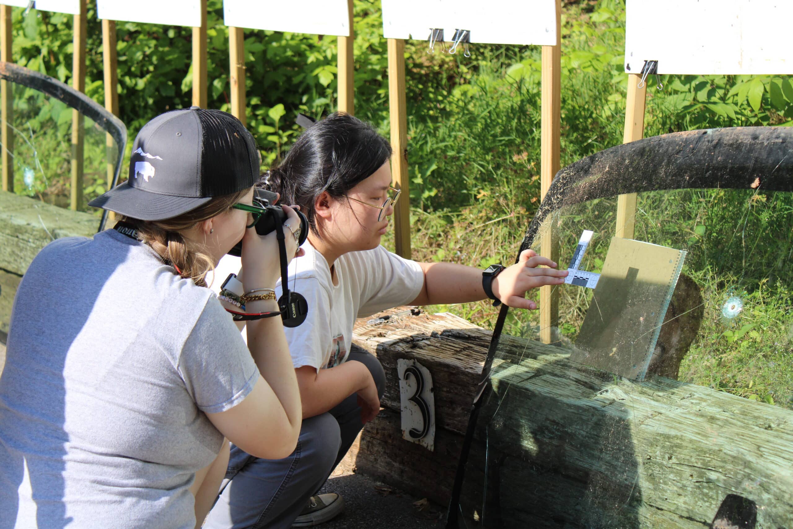 Two female students examine a windshield with a bullet hole in it. One student is measuring the hole and the other is photographing it.