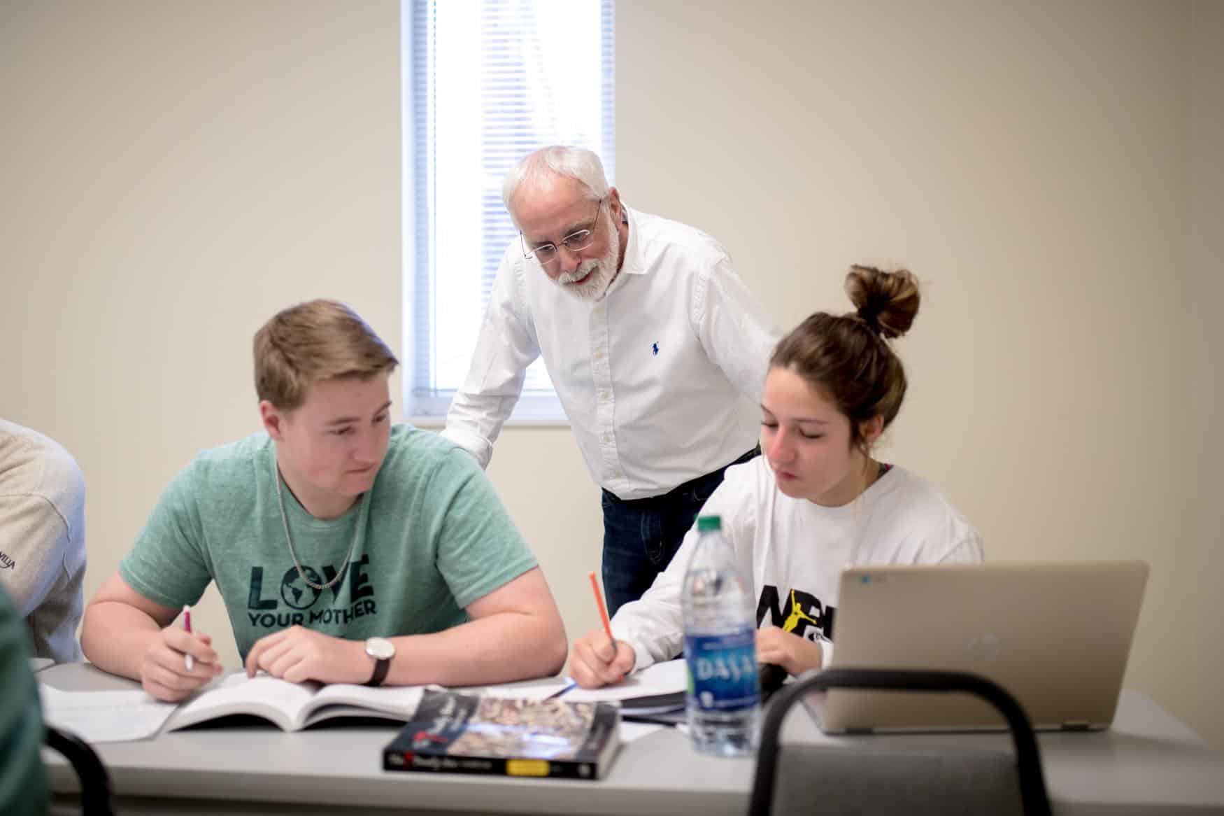 Professor in a white shirt leans over two students writing and studying.