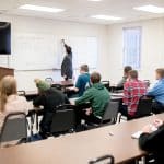 Class of students pays attention to a professor writing on a white board.