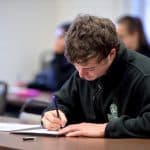 Young man looks down at a paper while writing at a desk.