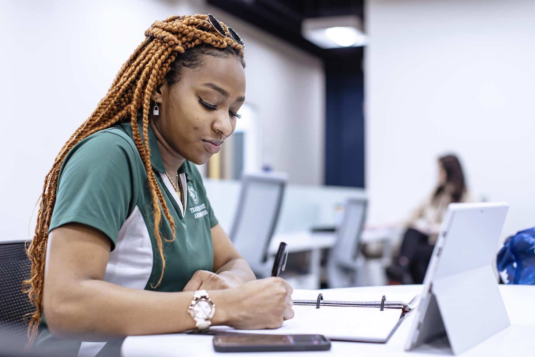 Young woman wearing a green Piedmont shirt takes notes at a table.