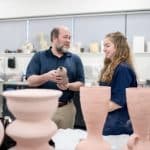 A professor holds ceramic pottery and speaks to a student beside him. The student is smiling and looking at the pottery.