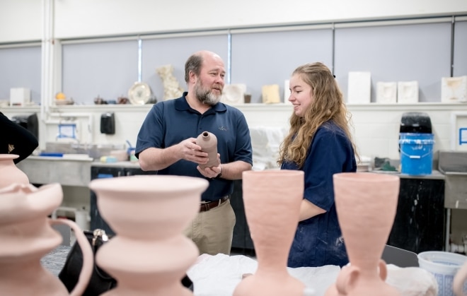 A professor holds ceramic pottery and speaks to a student beside him. The student is smiling and looking at the pottery.