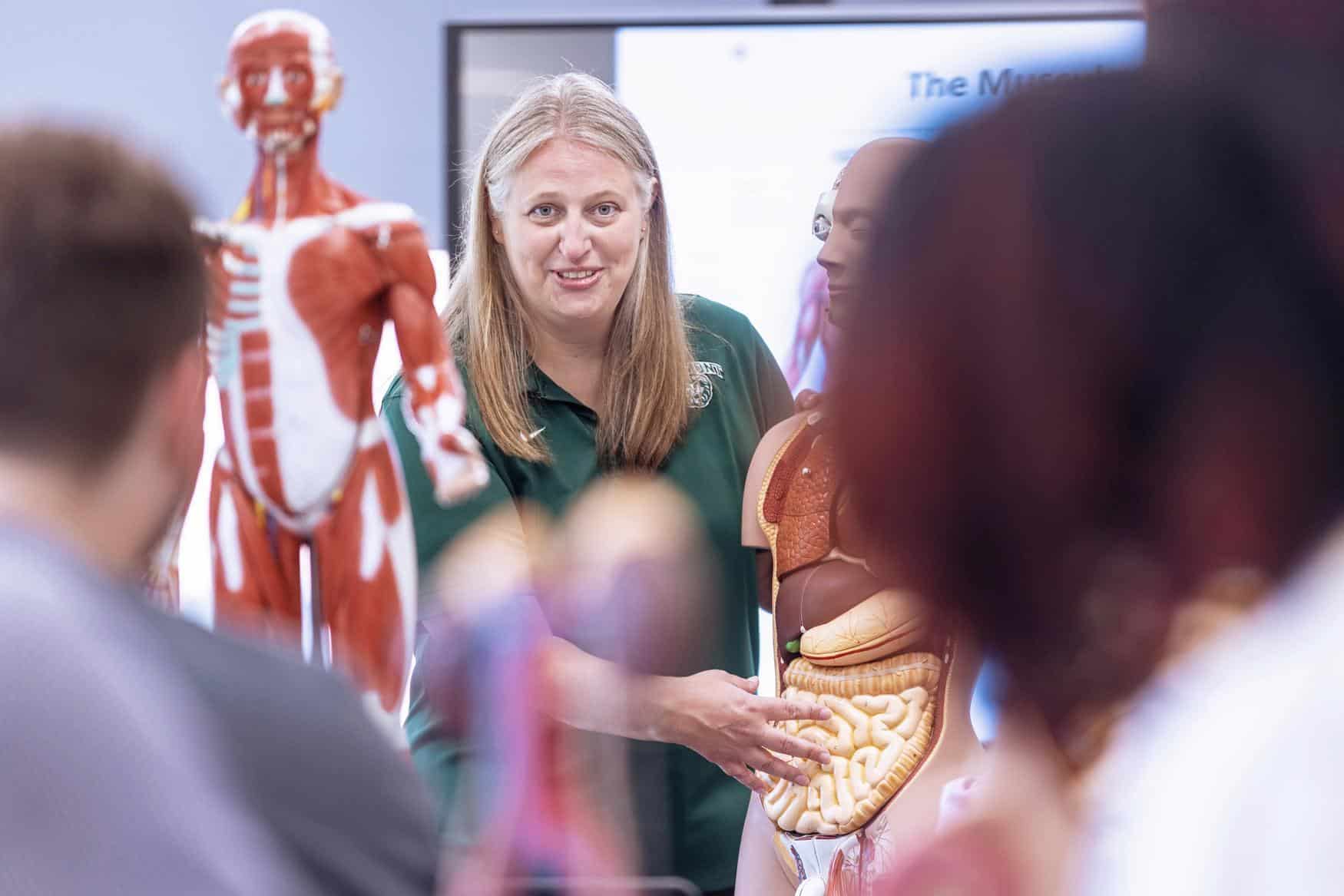 A woman demonstrates human anatomy using a life-sized model with exposed organs to a group of students in a classroom setting. Anatomical models and diagrams are visible in the background.