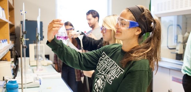 Young woman with safety glasses smiling and holding a glass beaker full of purple liquid