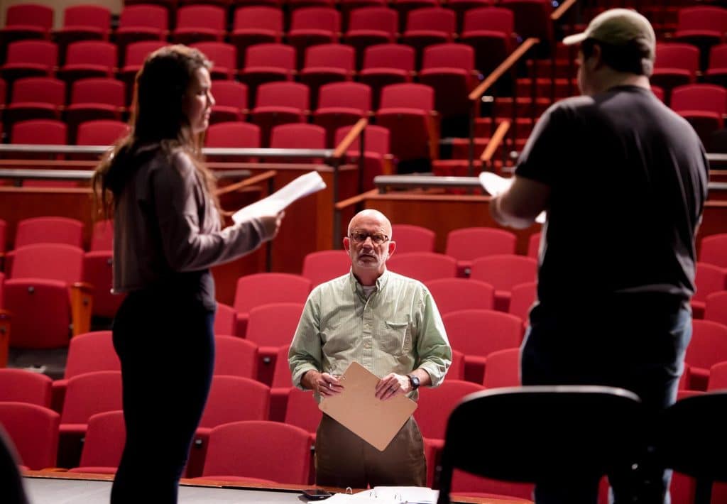 Drama professor looks towards the camera, framed by two students in silhouette acting out a scene. Red auditorium chairs are visible in the background.