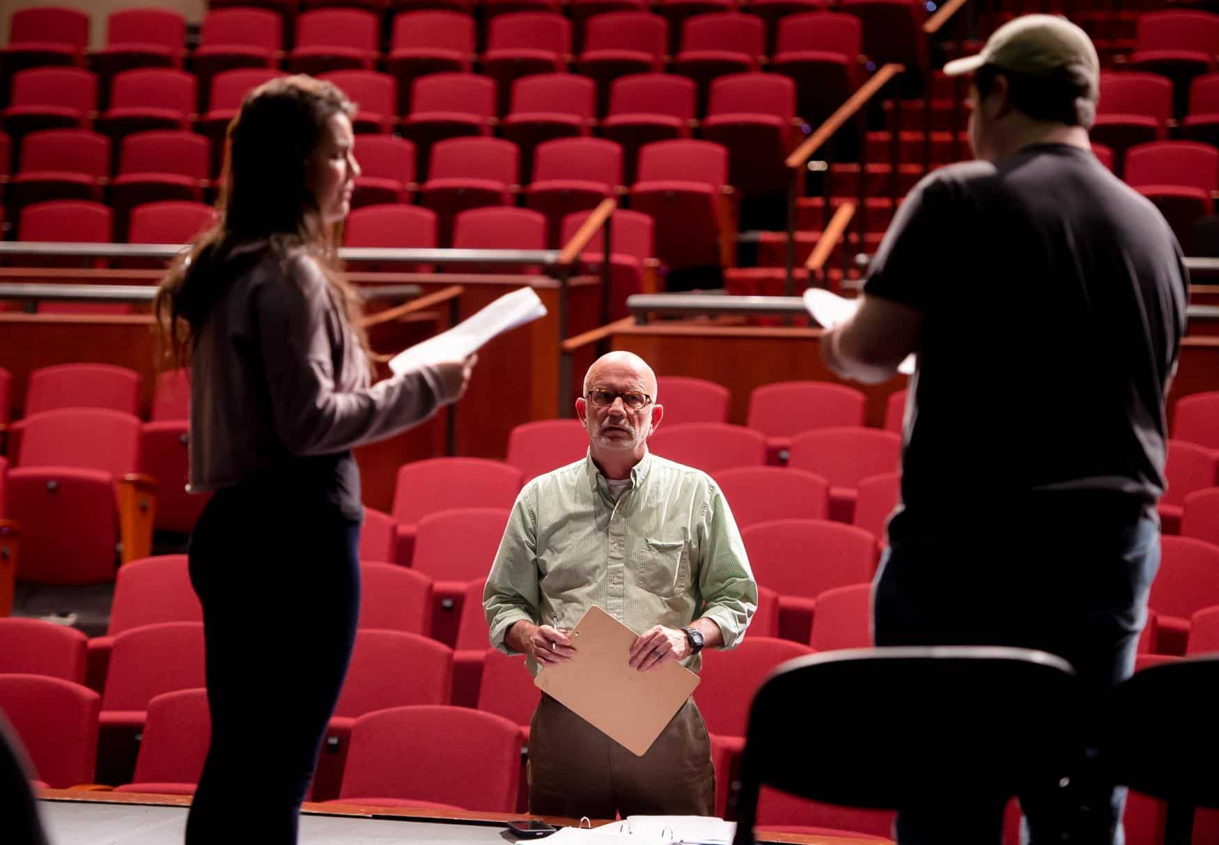Drama professor looks towards the camera, framed by two students in silhouette acting out a scene. Red auditorium chairs are visible in the background.