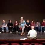 Student in a blue plaid shirt acting out a scene on a stage with fellow students and professor watching