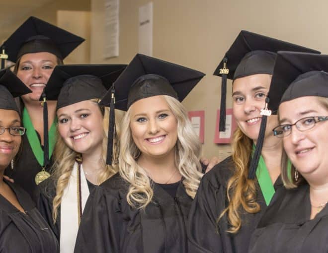 A group of eight women in black graduation gowns and caps, smiling and standing close together indoors, celebrating their graduation. Some wear green honor cords around their necks.