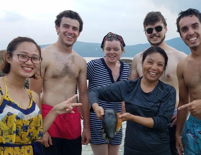 Six smiling people stand together in shallow water by rocks at the beach; one woman in a dark shirt is holding a fish, and others are pointing and laughing, with mountains visible in the background.