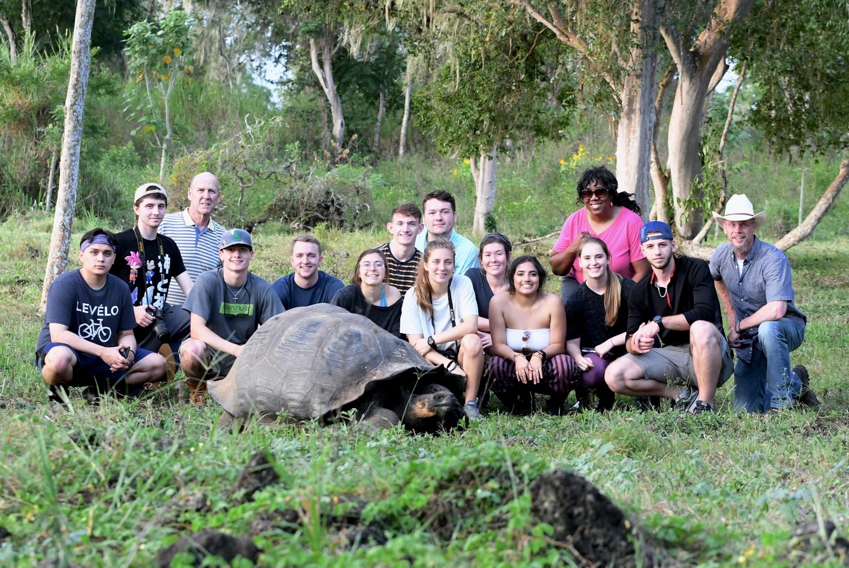 Group of students posing for an outdoor photo. In the foreground is a large tortoise and green grass. In the background are trees and tall grass.