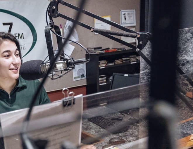 A person sits at a desk speaking into a microphone in a radio studio with a “Z98.7 FM Piedmont College” banner and audio equipment in the background.