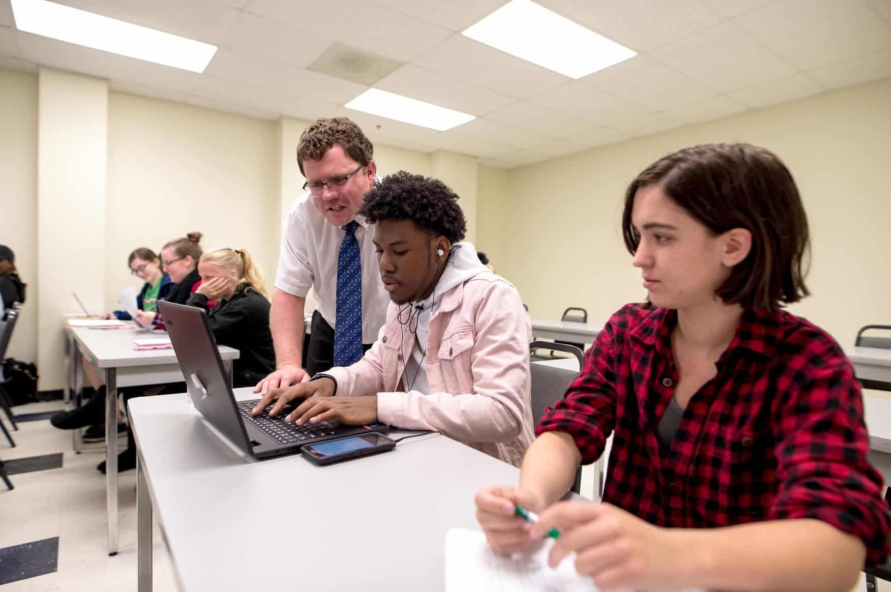 A teacher stands beside a student using a laptop in a classroom, offering guidance. Other students sit at desks, some taking notes and others listening attentively.