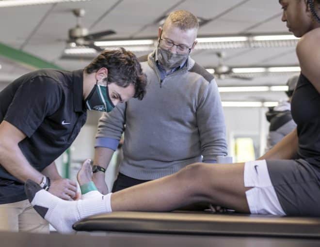A person wearing a mask tapes the ankle of a seated athlete on a treatment table, while another masked person observes in a clinical or athletic training setting.