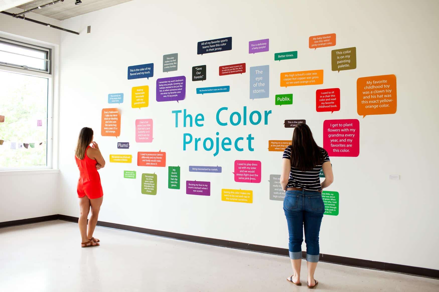 Two women stand in a bright gallery, reading colorful text boxes on a wall titled The Color Project, displaying various people’s quotes about their favorite colors and personal experiences with color.