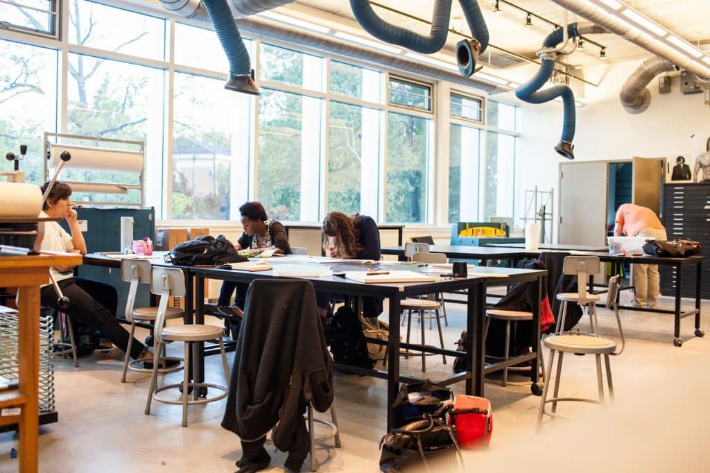 Several students work and study at large tables in a bright, modern classroom or studio with tall windows, stools, art supplies, and overhead ventilation ducts. Some are seated and one person is standing near a cabinet.
