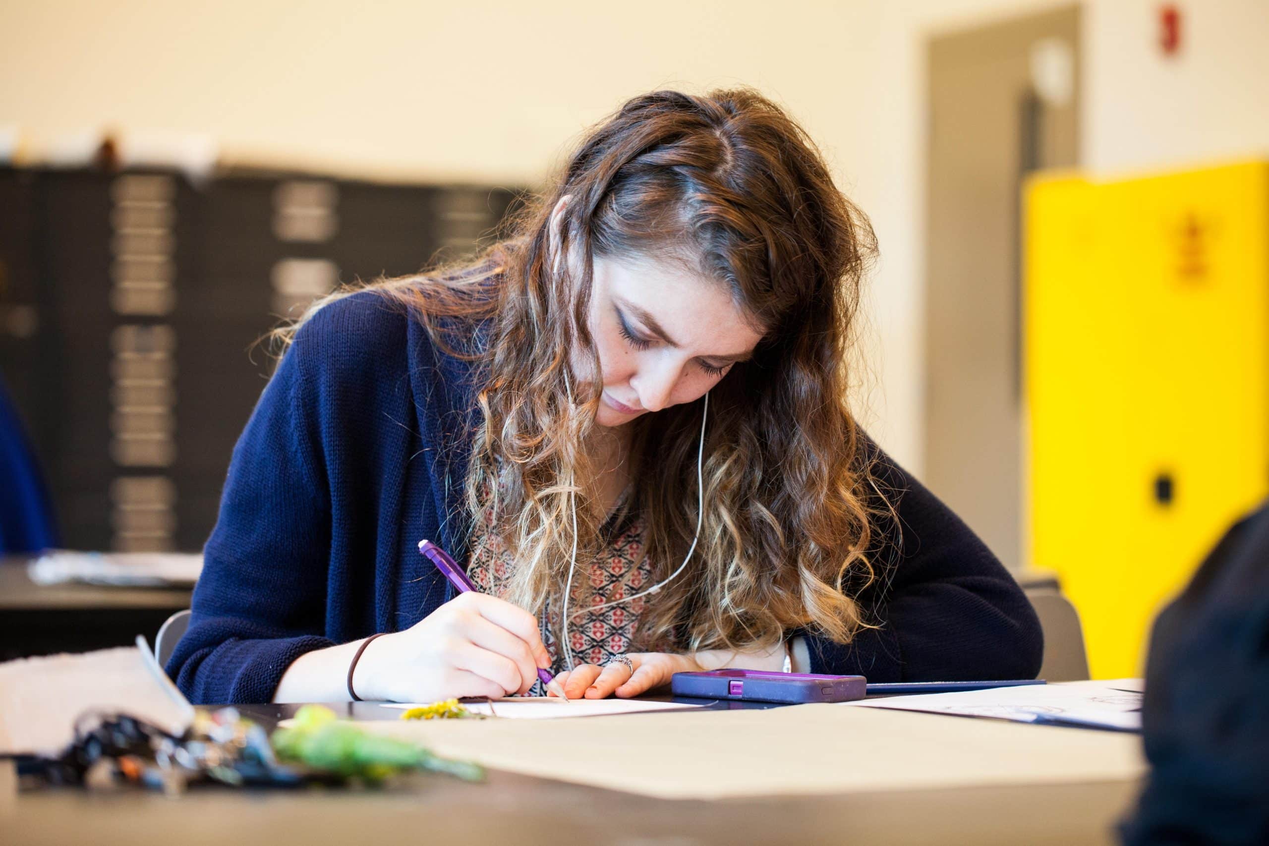 A young woman with long wavy hair writes on paper at a desk, focused on her work. She wears earbuds and a dark cardigan, with a calculator, papers, and small objects on the table in front of her.