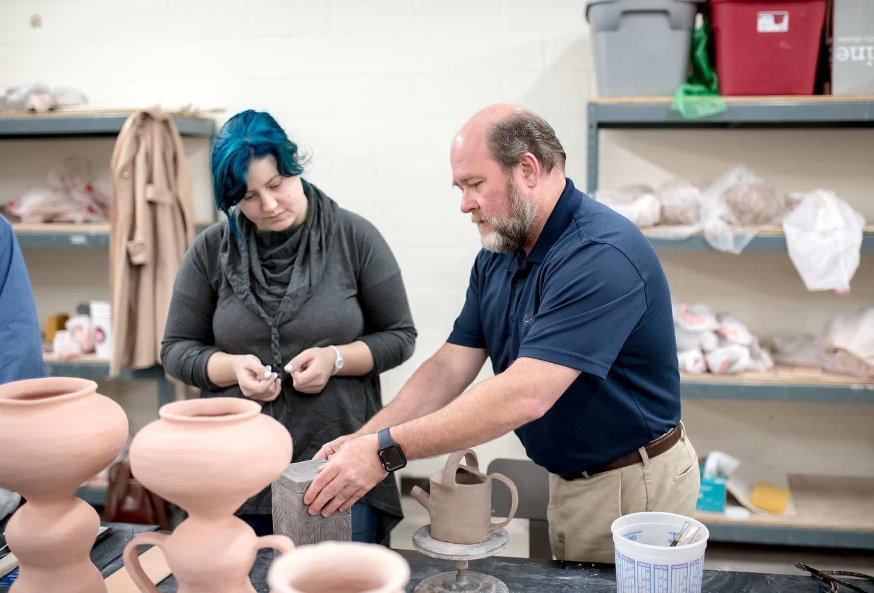 A man demonstrates pottery techniques to a woman with blue hair in a ceramics classroom, surrounded by unfinished clay pots and art supplies on a table. Shelves with containers and materials are visible in the background.