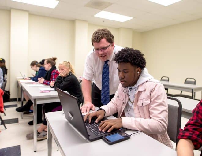 Professor wearing a white button down shirt and blue tie, leaning over a young man in class. Both are looking at a laptop in front of the young man.