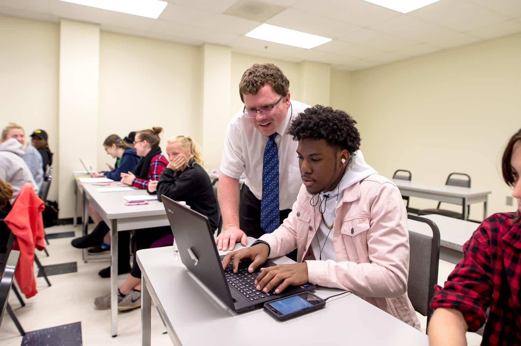 Professor wearing a white button down shirt and blue tie, leaning over a young man in class. Both are looking at a laptop in front of the young man.