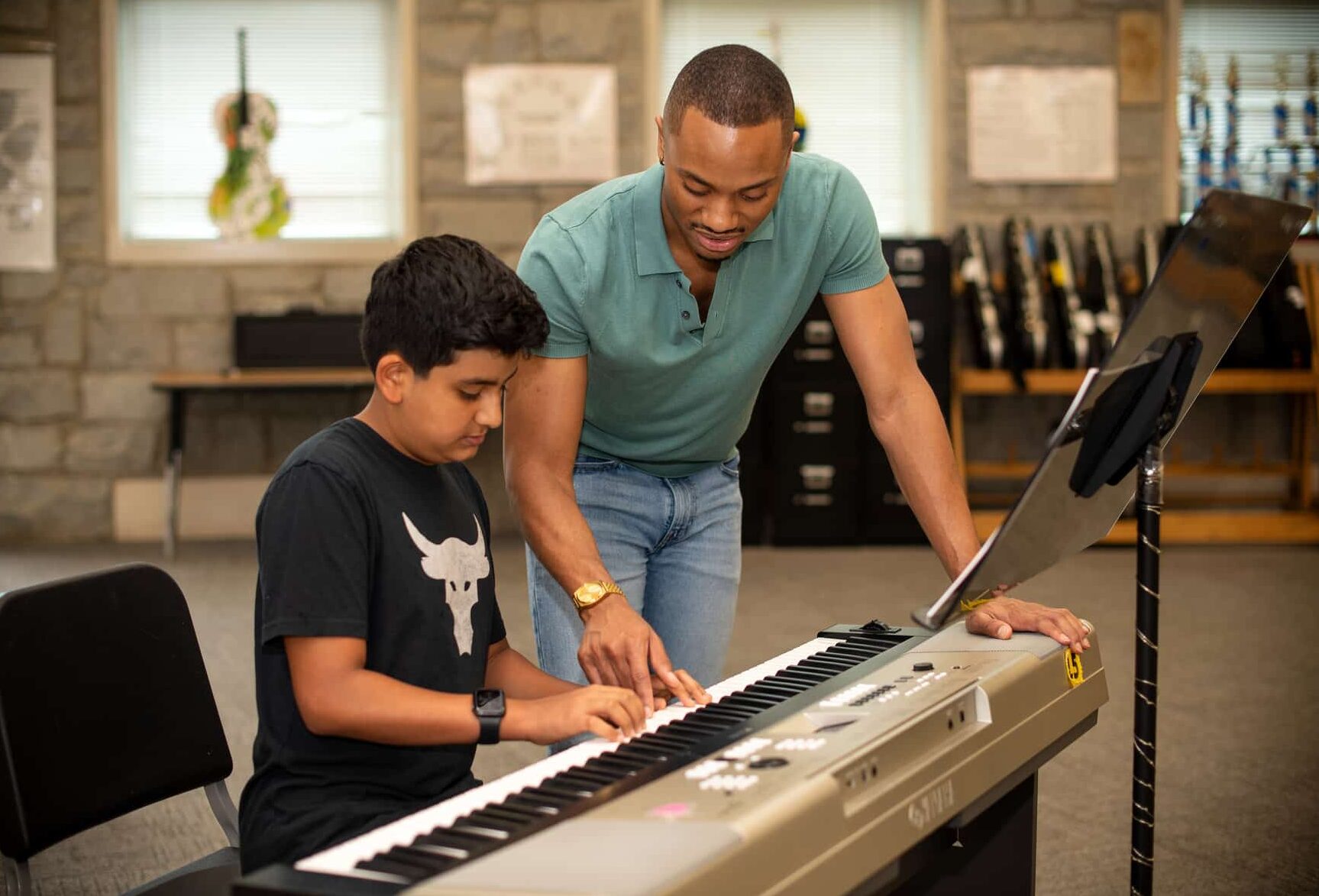 Young man leaning over an electronic keyboard, teaching a young boy chords.