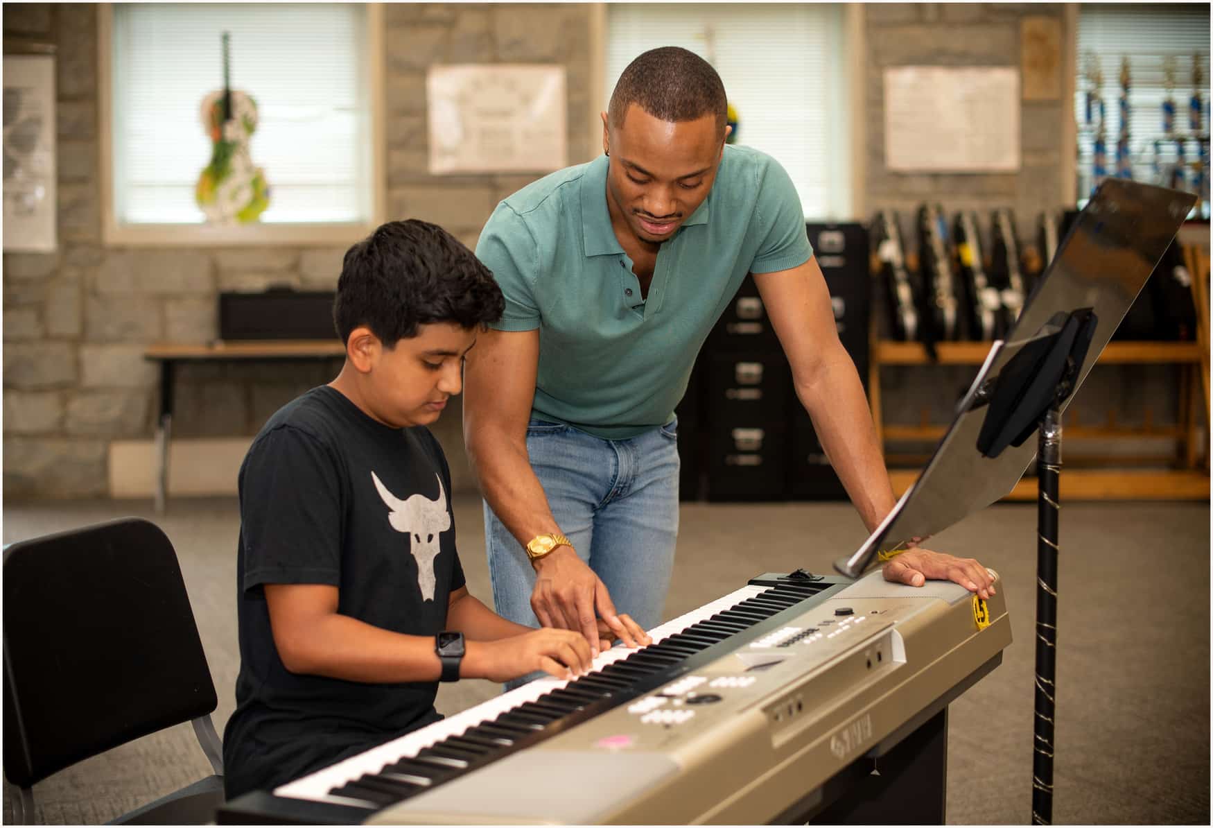 Young man leaning over an electronic keyboard, teaching a young boy chords.