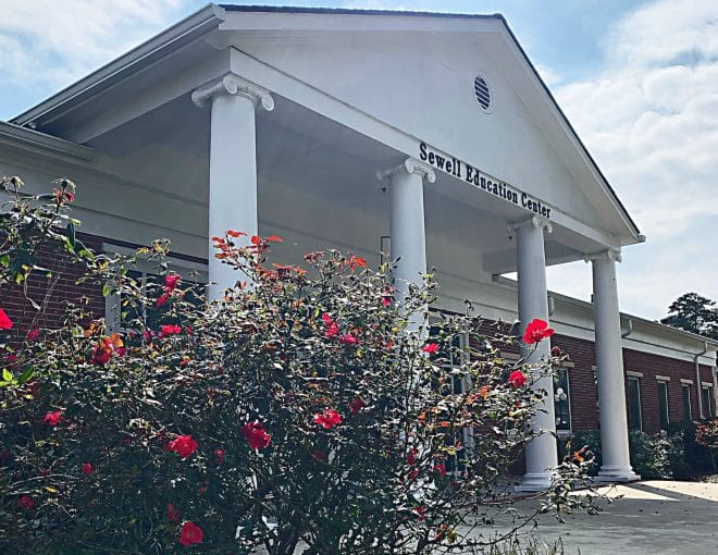 The Sewell Education Center building with white columns and a triangular pediment, partially obscured by blooming red flowers and green shrubs under a partly cloudy sky.