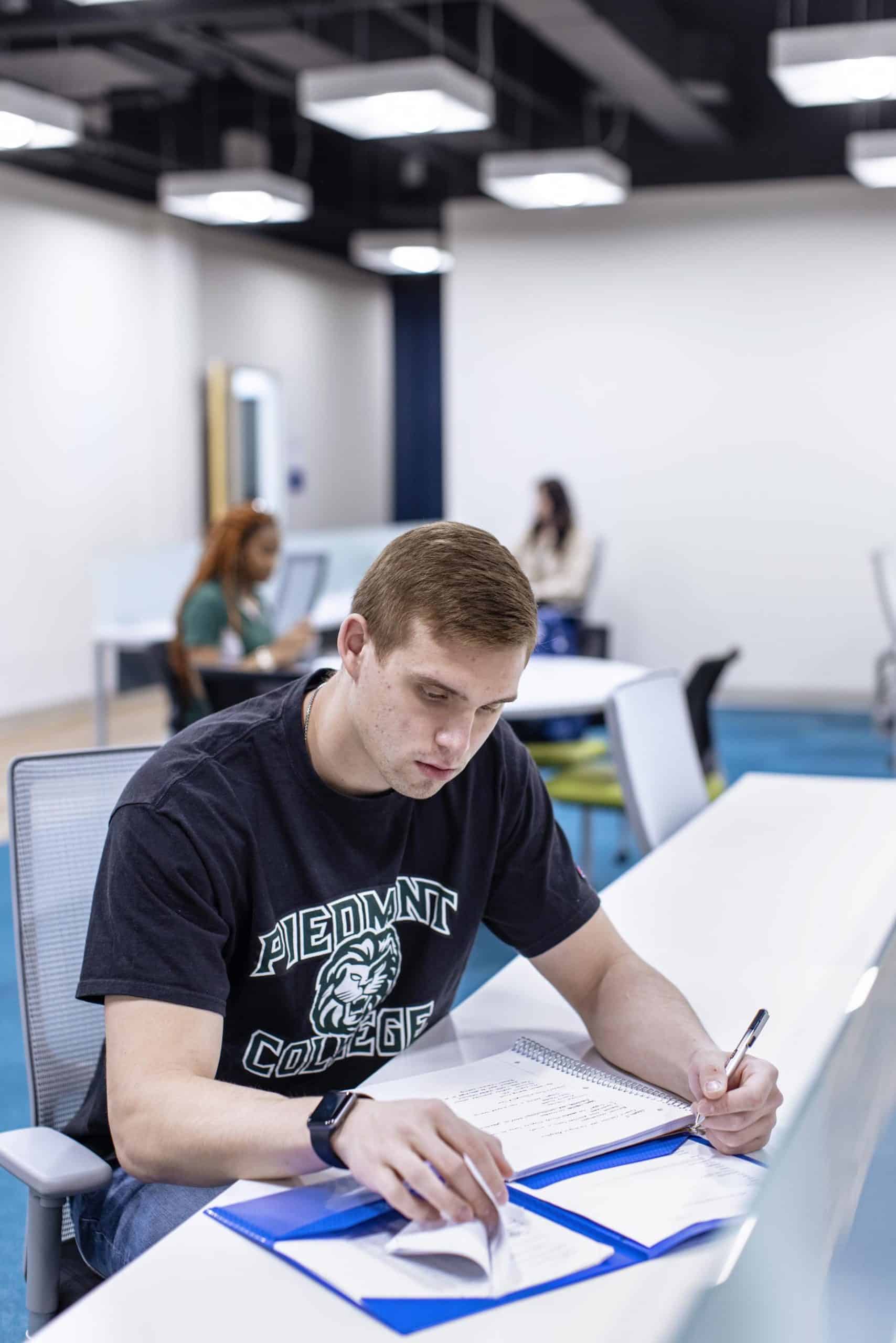 A young man wearing a “Piedmont College” t-shirt writes in a notebook at a desk in a modern study area. Two other students are studying in the background at separate tables.